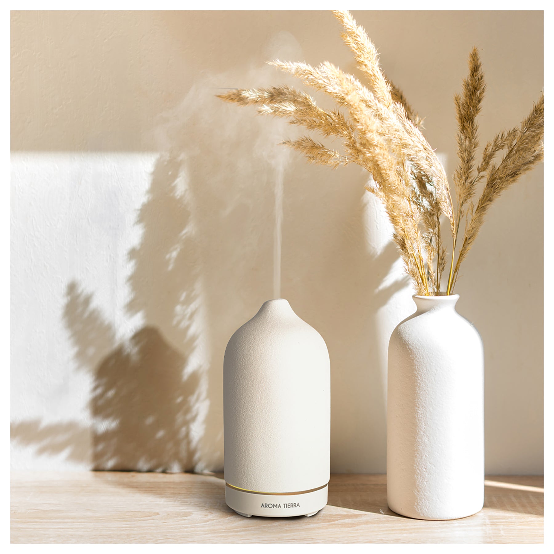 A white ceramic electric diffuser emitting steam, placed on a wooden surface with a vase of dried plants in the background.