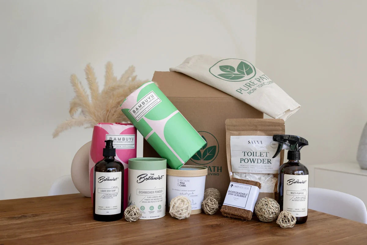 Non-toxic home cleaning bundle from Pure Path featuring The Botanist cleaners, Sanna toilet powder, Bambuyu bamboo paper, and eco-friendly dish scrubbers on a wooden table.